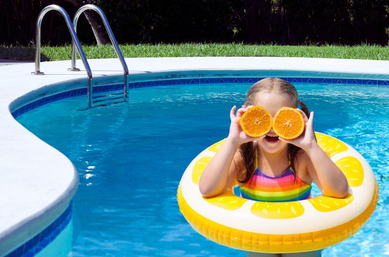 A joyful little girl wearing a rainbow swimsuit, standing in a pool and holding oranges with a big smile on her face
